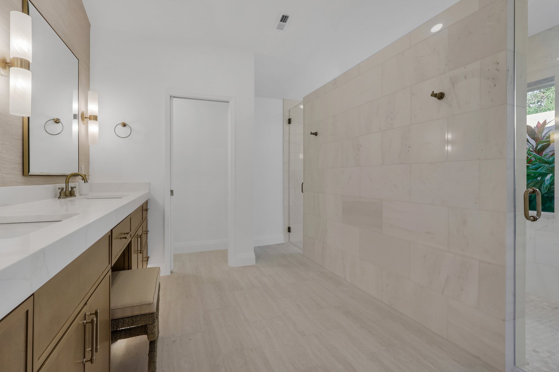 Primary bath with a warm oak vanity, aged brass fittings and a stone shower wall, designed by Iglesias Design Group
